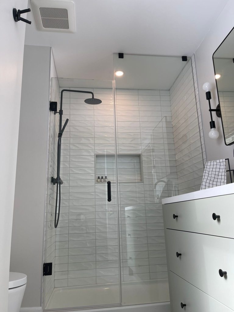 Markham bathroom remodel featuring a sleek walk-in shower with matte black rain showerhead, frameless glass door, white textured wall tiles, and a modern white vanity with black handles.