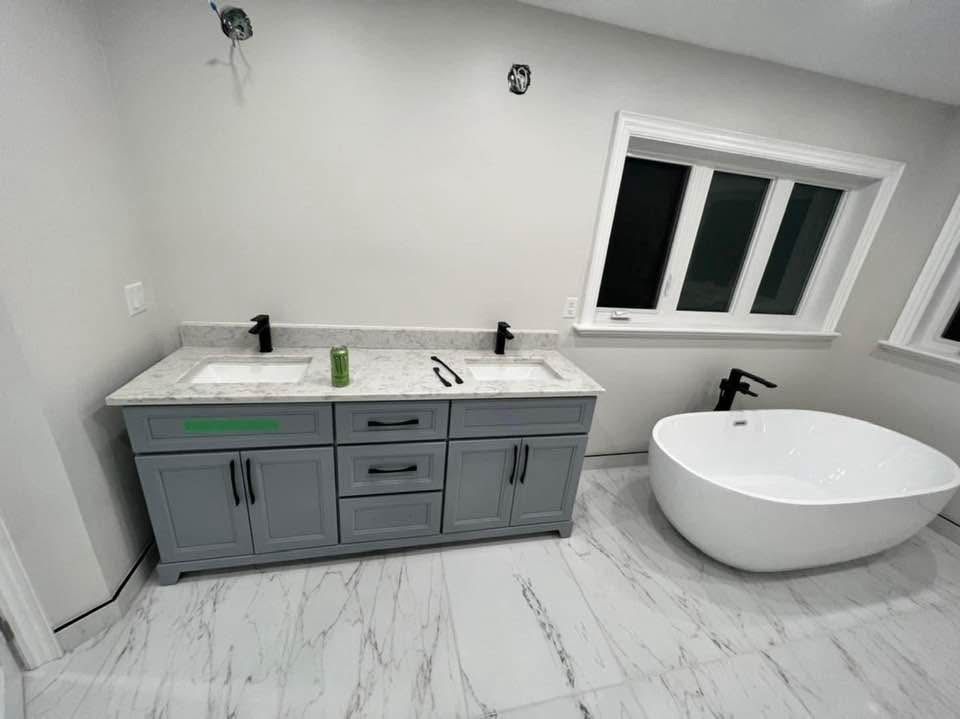 Grey shaker-style double sink vanity with white marble countertop and matte black faucets beside a freestanding white bathtub on marble-patterned tile flooring