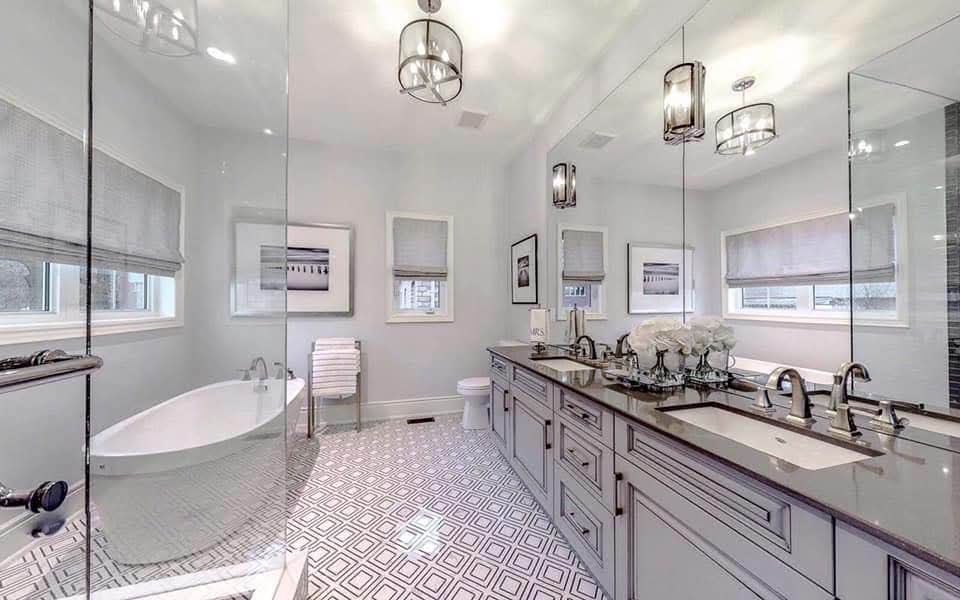 Luxury Scarborough master bathroom with a grey double-sink vanity, freestanding white bathtub, patterned geometric tile flooring, large mirrors, and modern pendant lighting.