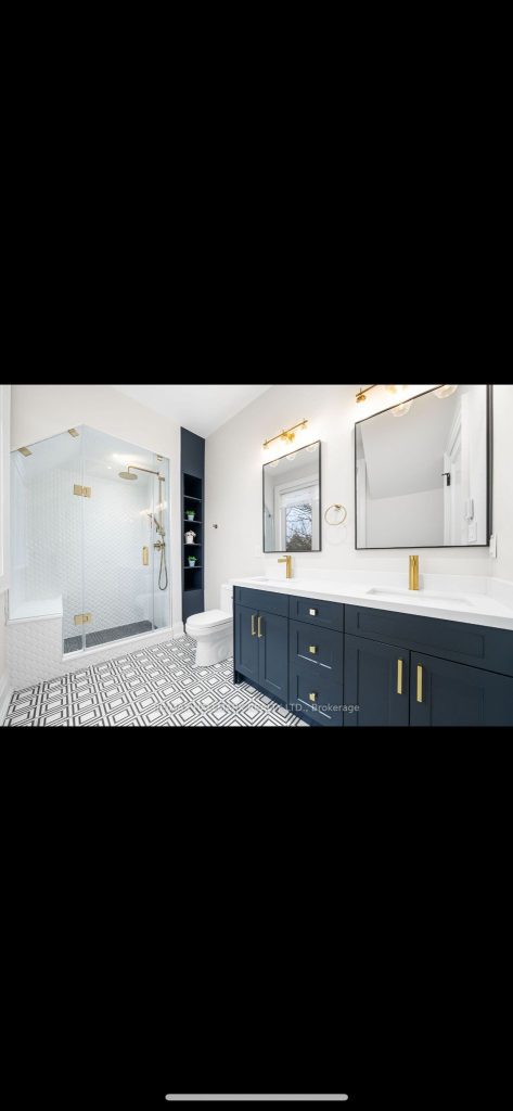 Modern bathroom with a navy blue shaker-style double sink vanity topped with white quartz, gold hardware and faucets, geometric black-and-white floor tile, dual mirrors with gold sconces, and a frameless glass shower enclosure.